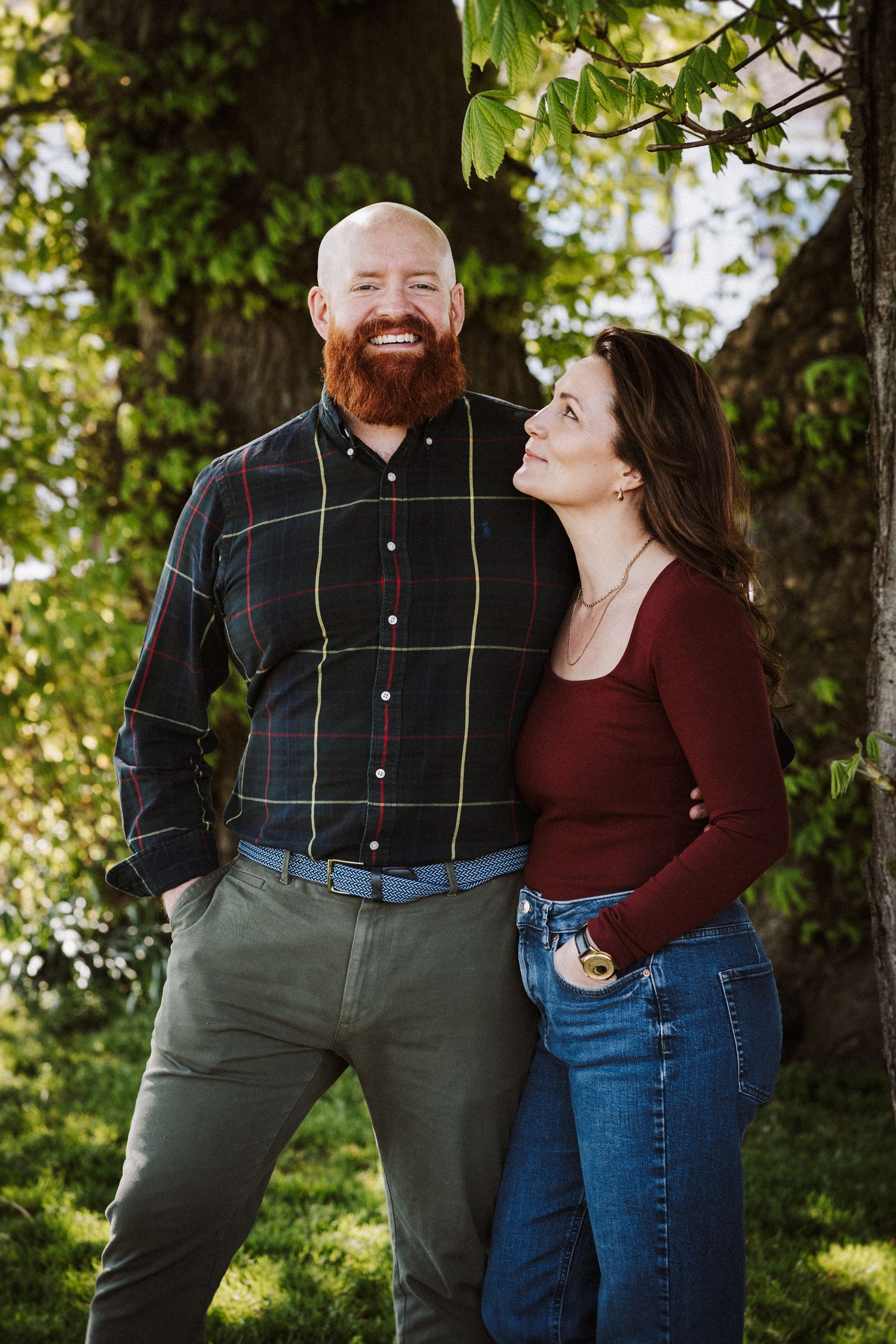 A couple laughs together while posing for photos in a park setting with trees in the background.