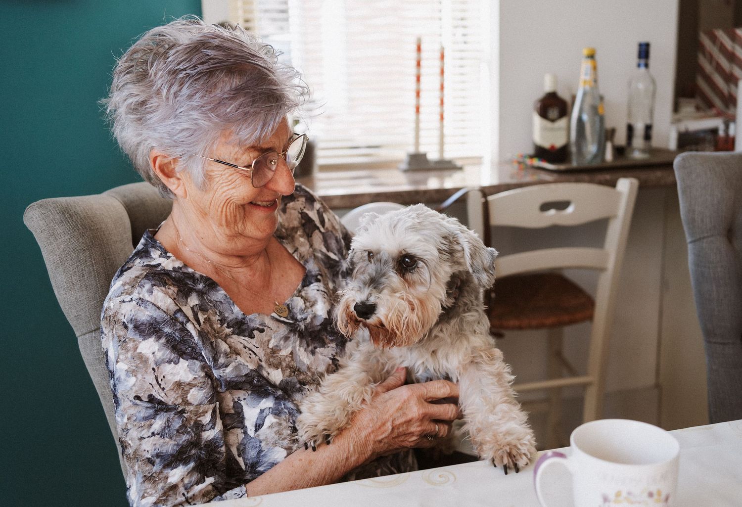 A small white dog sits with someone in a floral top at a kitchen table with coffee cups.