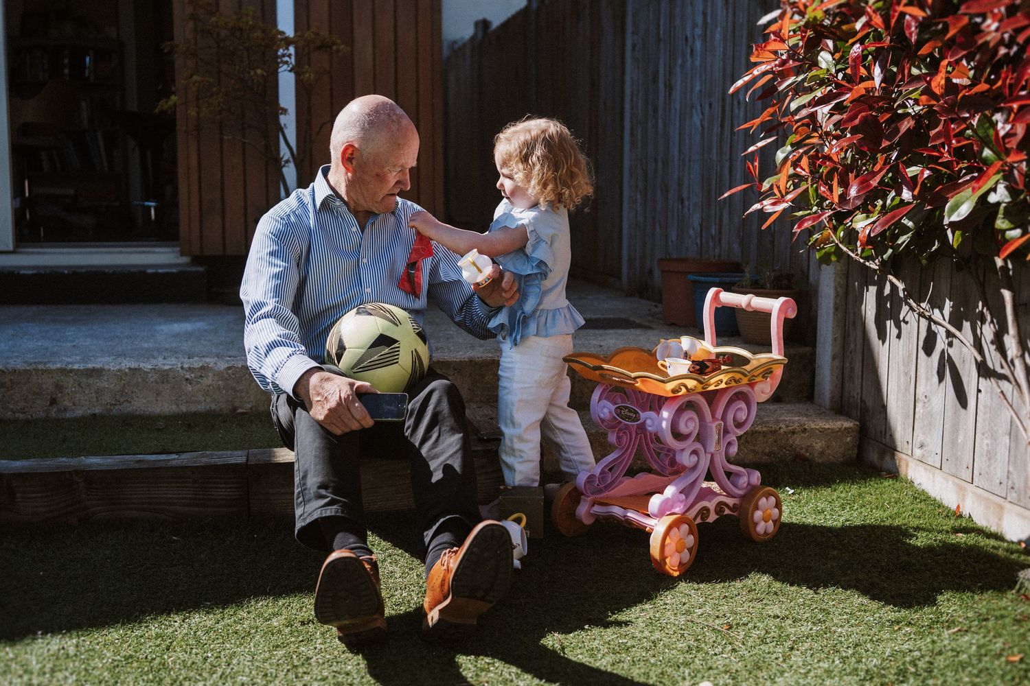 A heartwarming outdoor scene with a child and adult sitting on grass with toys and a pink stroller on a sunny day.