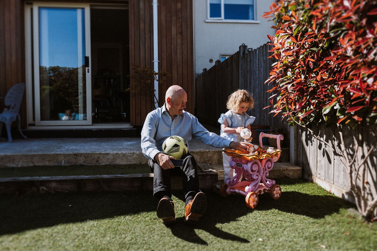 A family relaxes together on a sunny lawn outside their home with red foliage visible in the background.