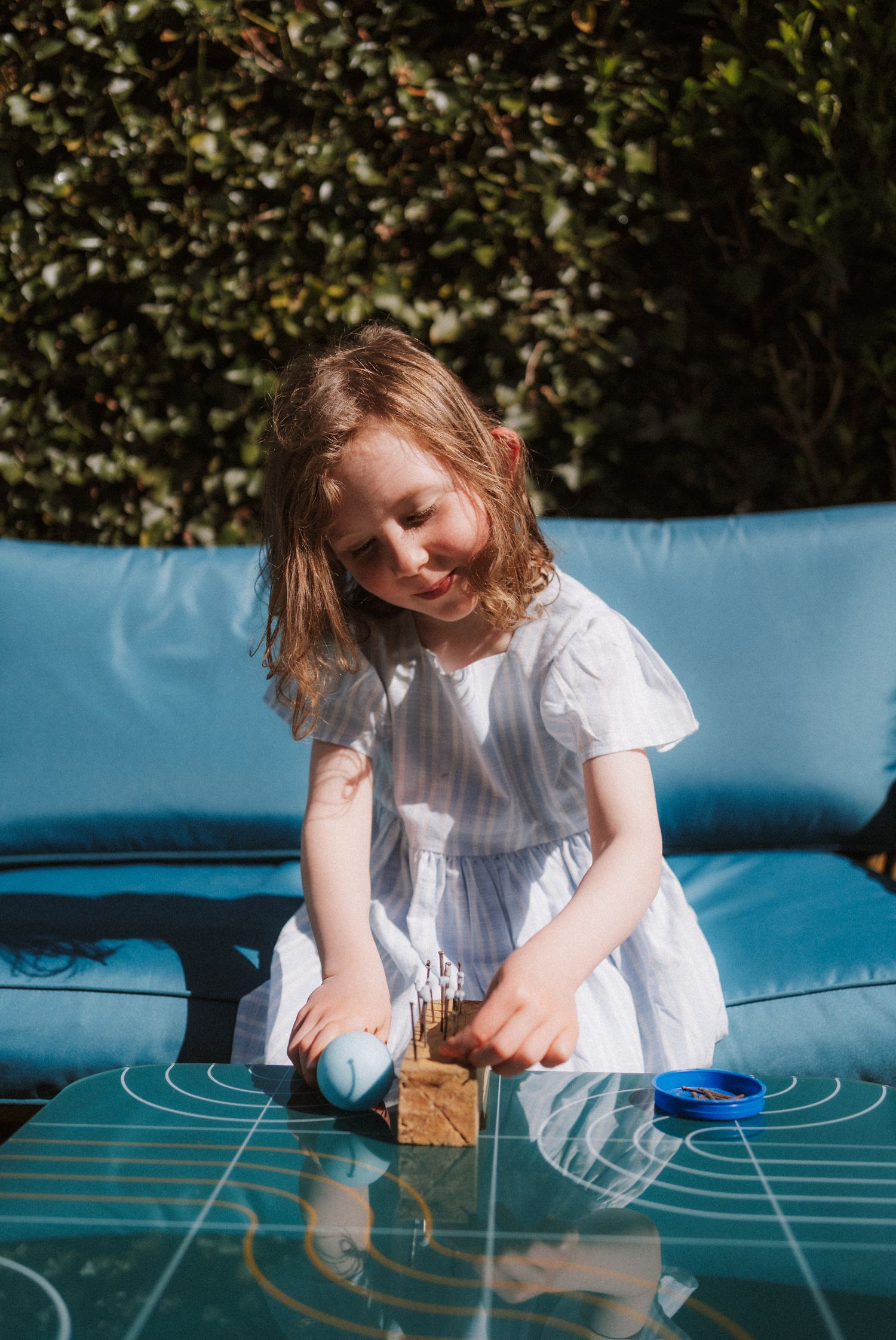 A child in a white dress plays with a blue chalk drawing on a patio table on a sunny day.