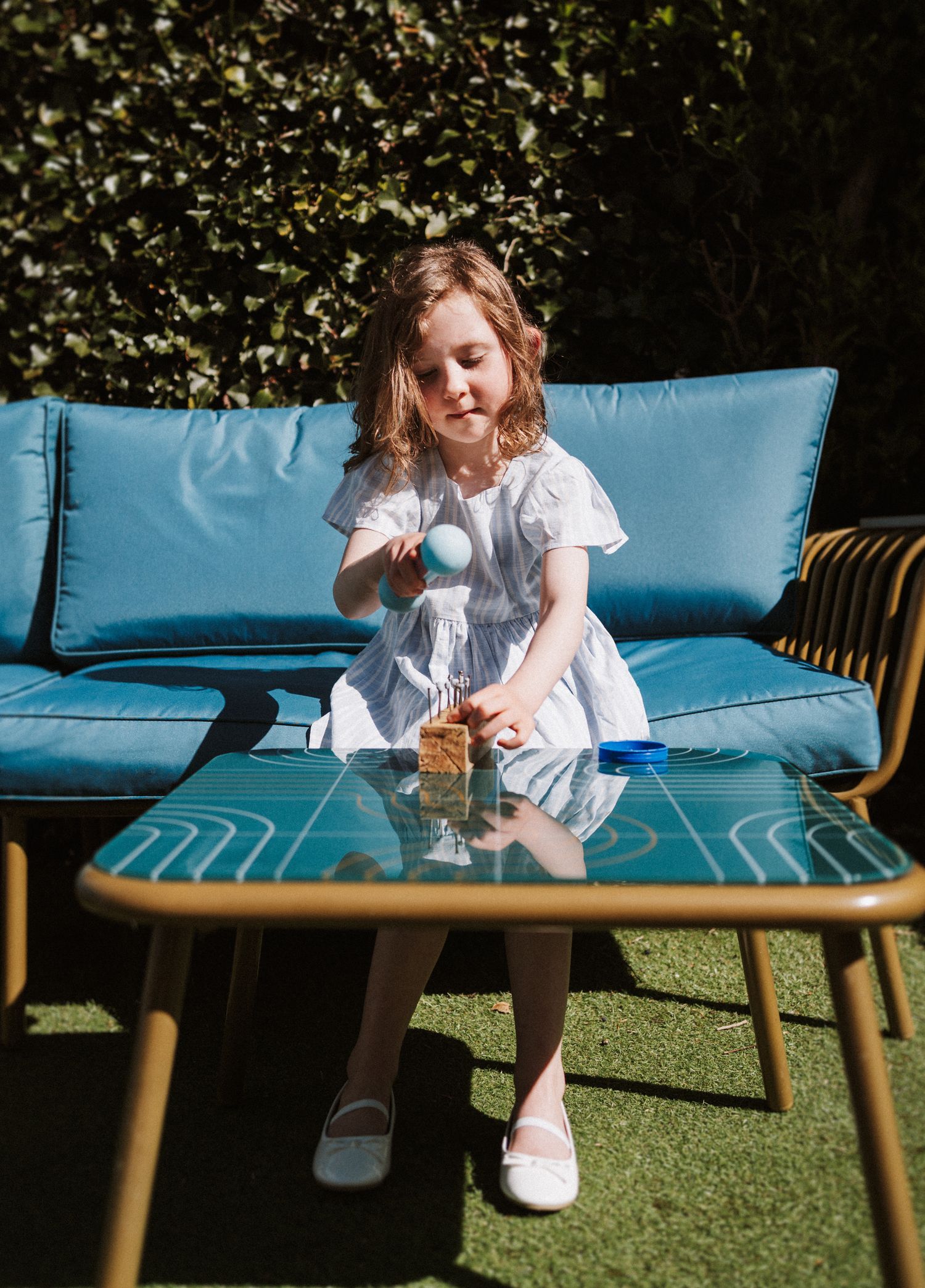 A child in a white dress sits on a blue outdoor sofa cushion against artificial turf in warm sunlight.