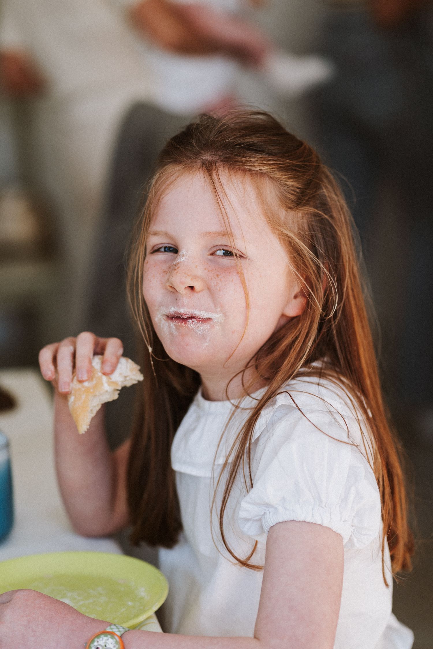 A young child in a white shirt takes a bite of food while sitting at a table.