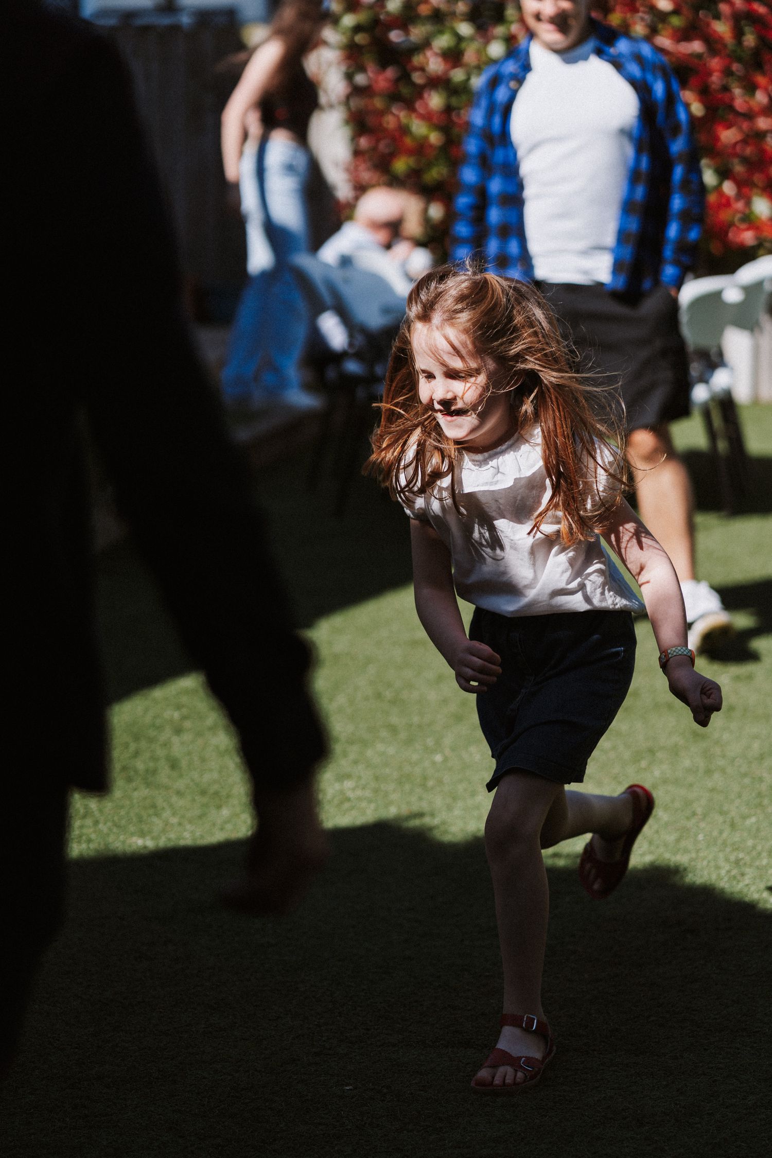 A child in a white top and dark shorts runs joyfully across green grass on a sunny day with hair flowing in the breeze.