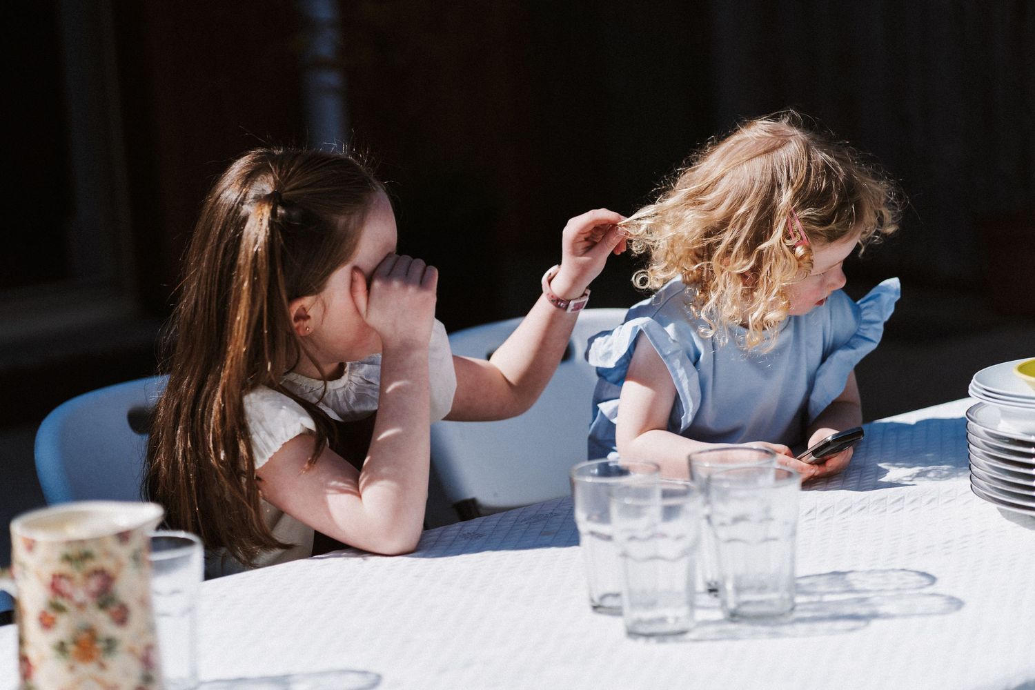 Two children sitting at a dining table with glasses and dishes, one resting head on hand while the other looks down.