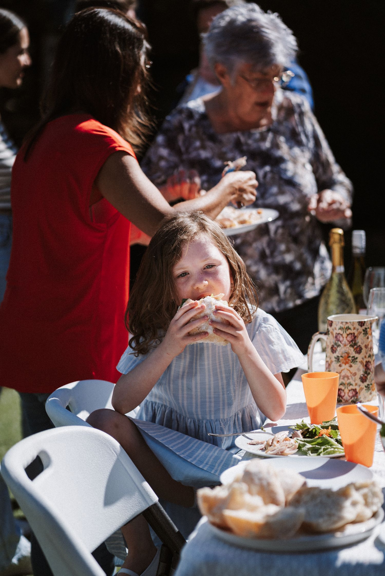 People enjoying a casual outdoor meal together at white folding tables on a sunny afternoon.