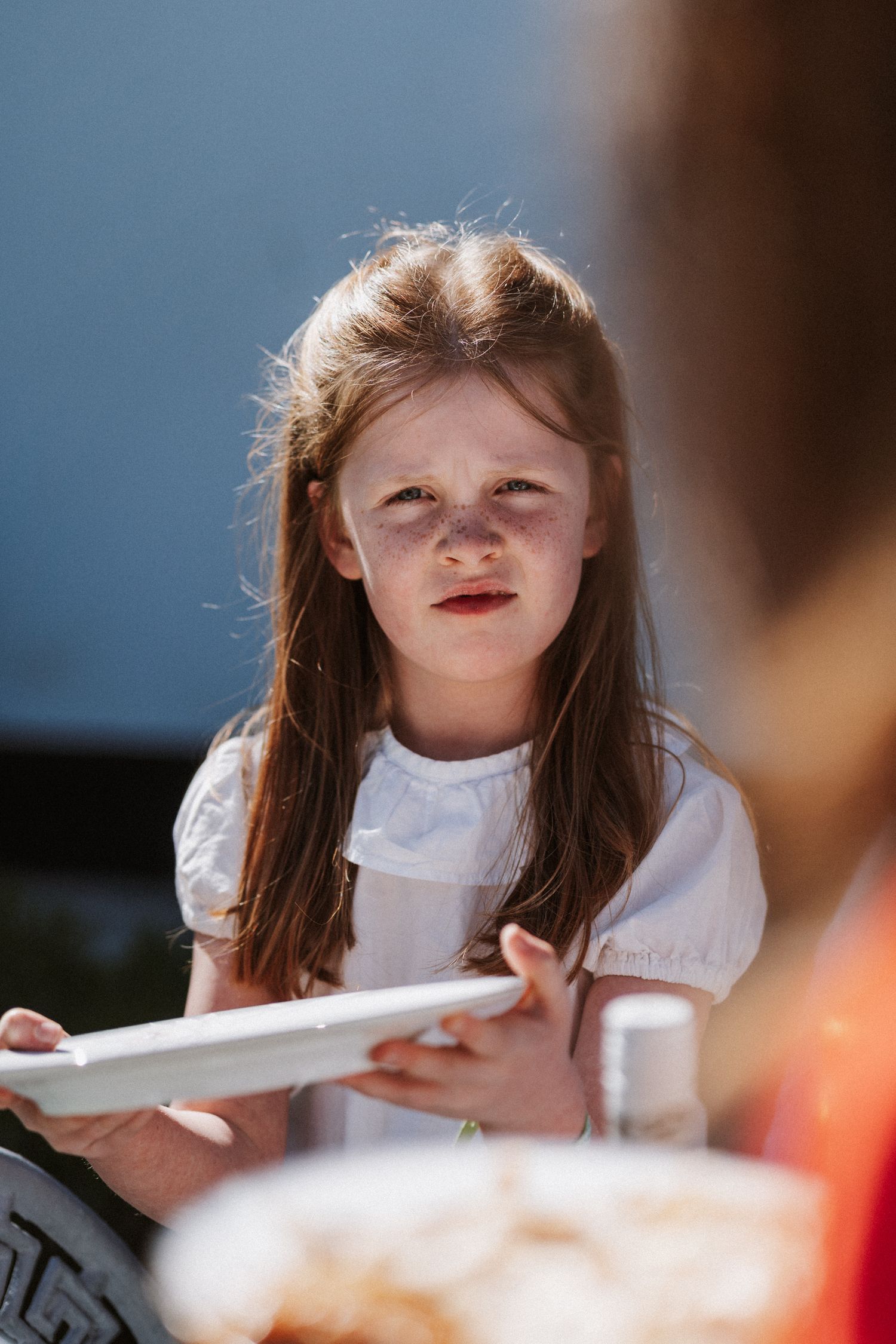 A child in a white dress leans forward at a table with a thoughtful expression in a sunlit setting.