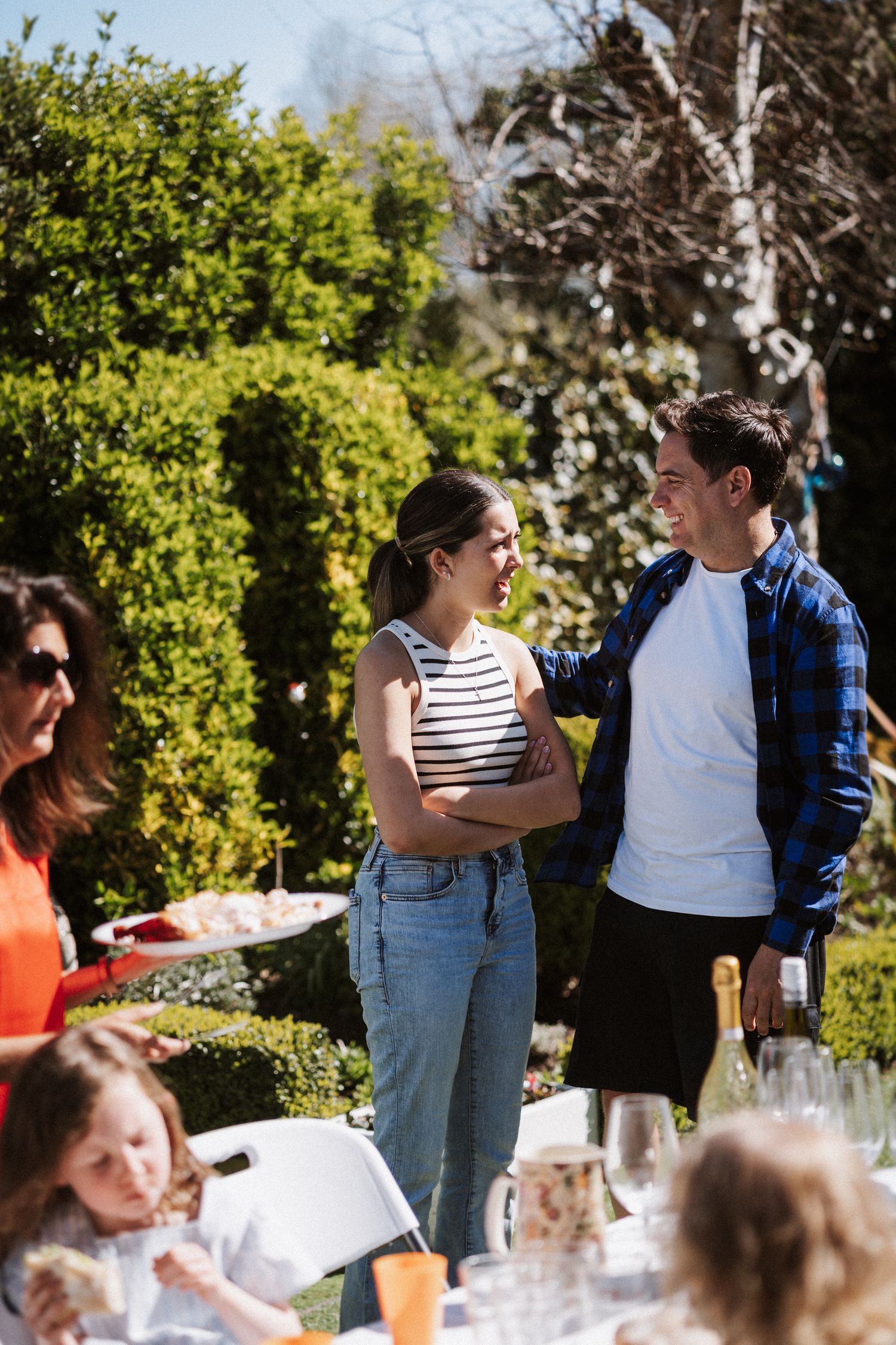 People gathered outdoors at a casual daytime event with tables and greenery in the background.