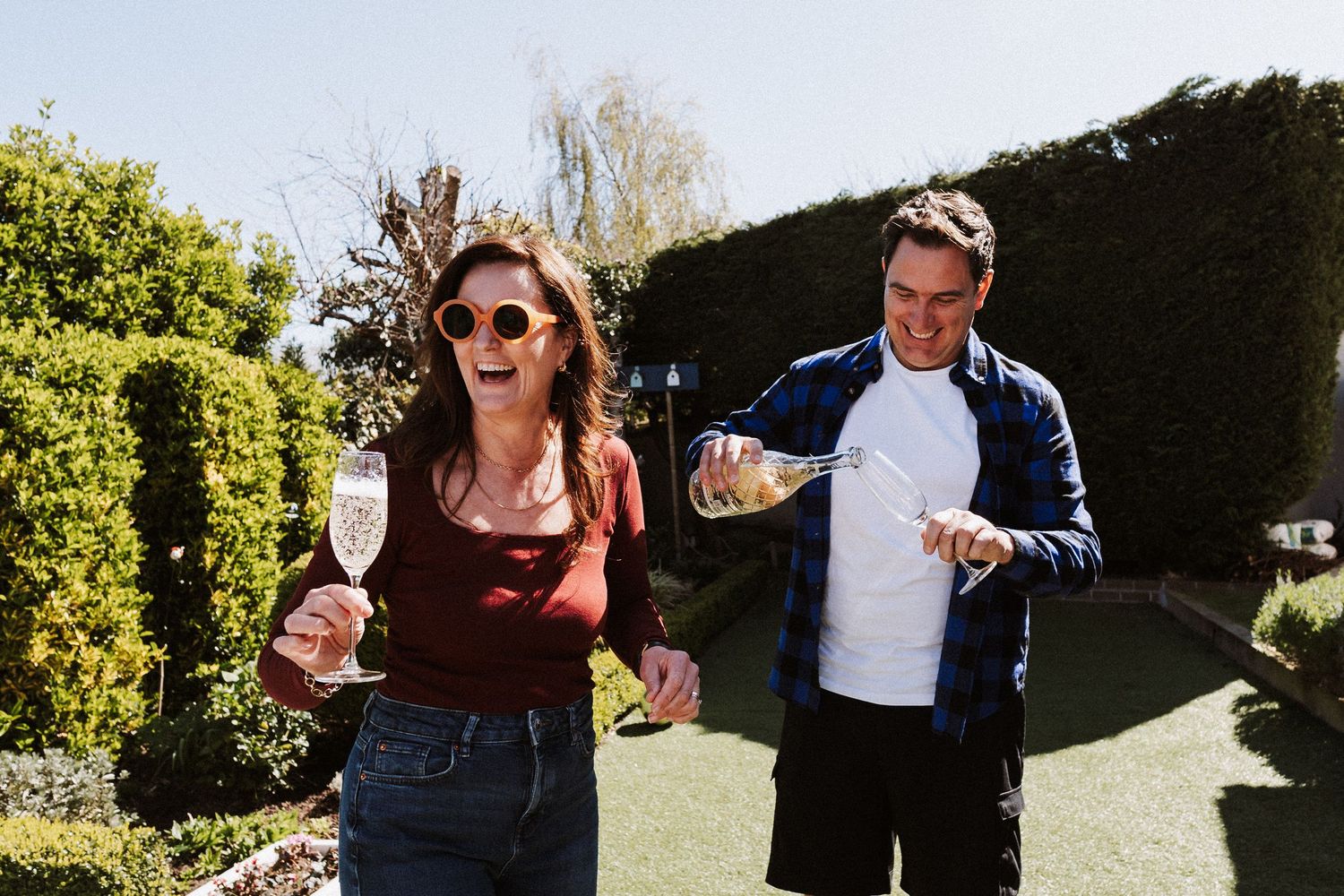 People dancing together in a garden with green hedges and trees in the background.