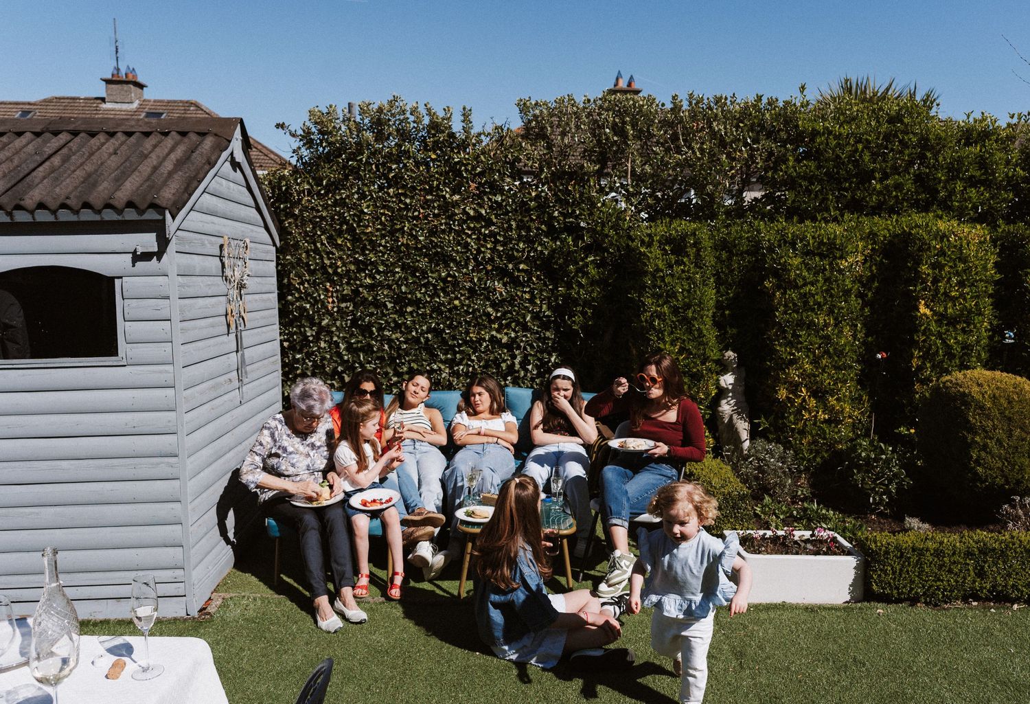 People enjoying a sunny outdoor gathering in a backyard with white garden buildings and hedges in the background.