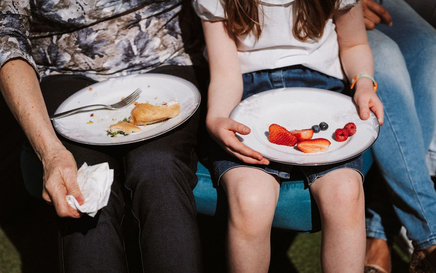 A row of people sitting together outdoors holding white plates with food and drinks during a casual gathering.