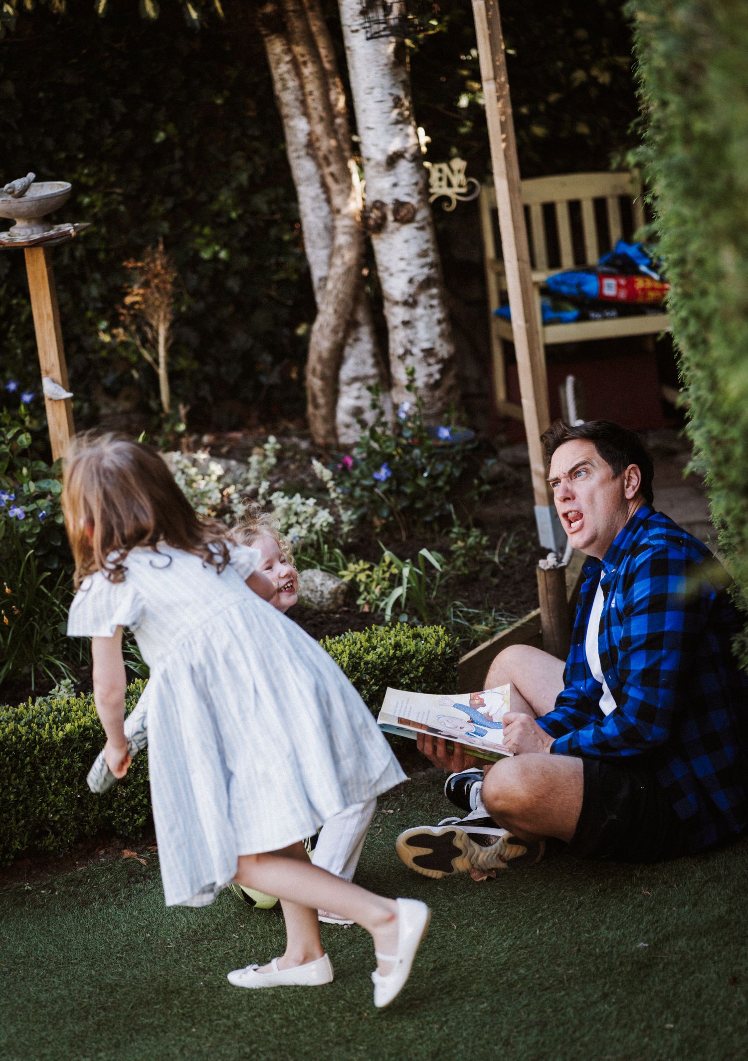 A couple sits together outdoors on grass near birch trees while sharing a playful moment in casual attire.