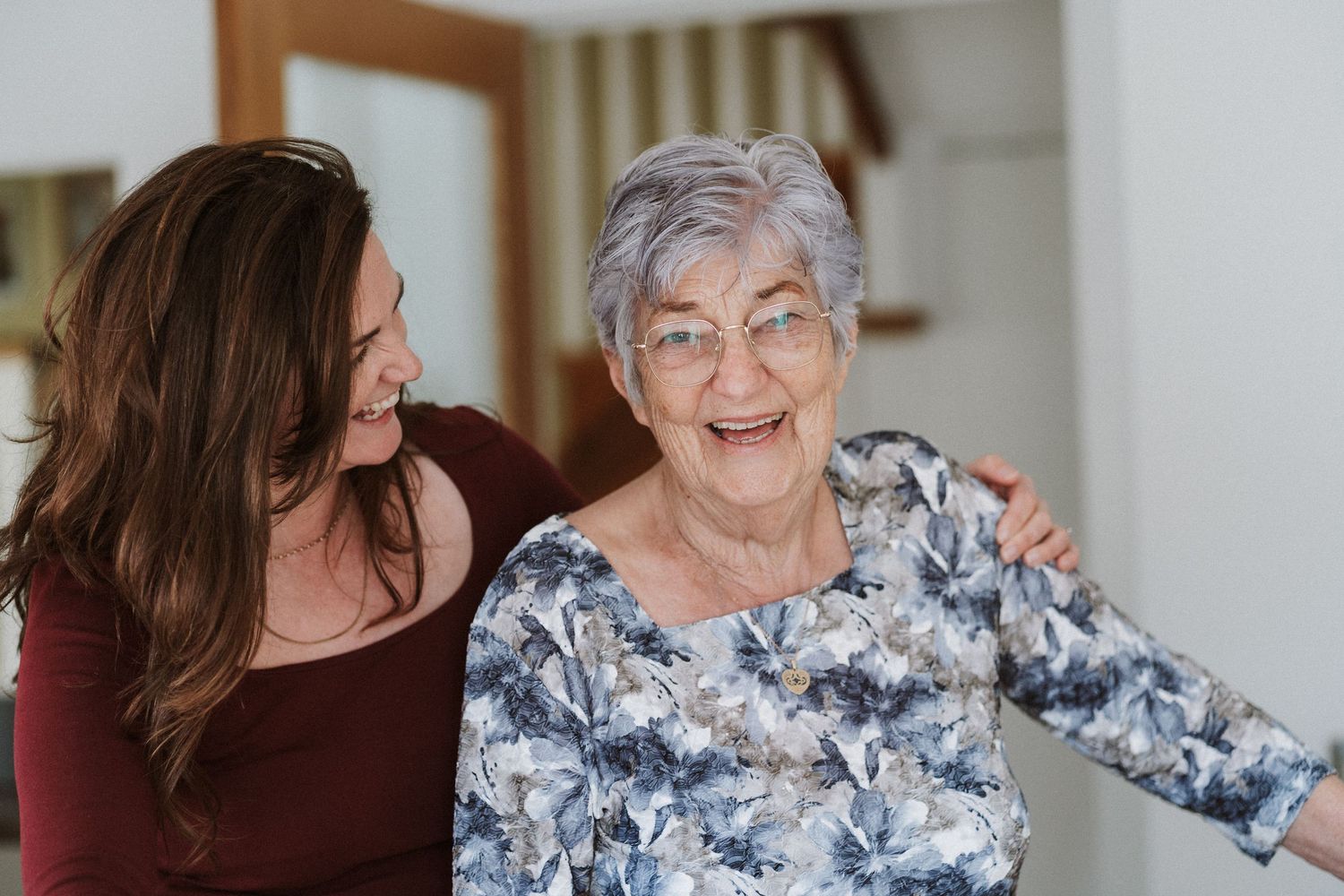 Elderly woman in floral dress and caregiver share joyful moments together in a bright indoor setting.