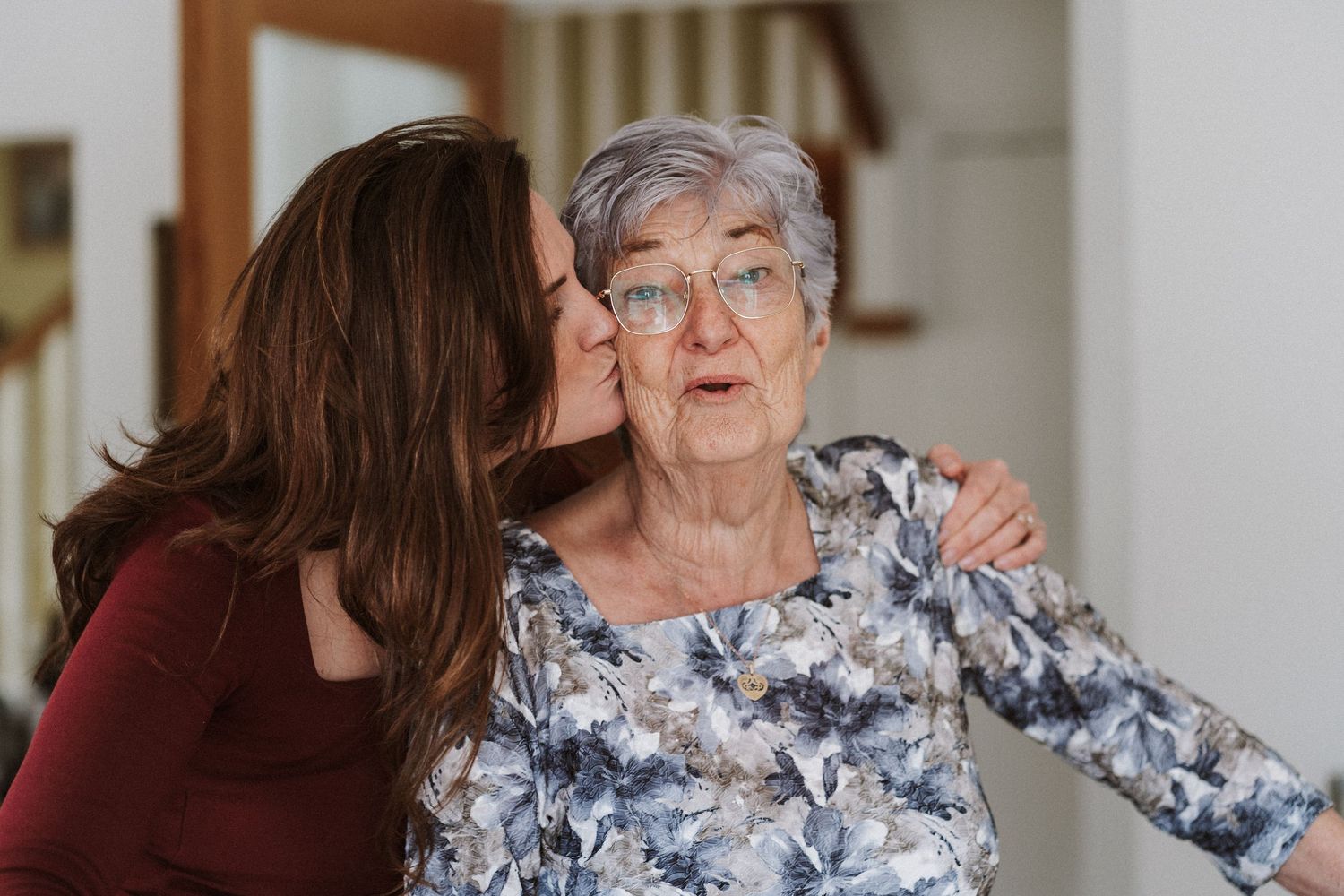 A senior woman and caregiver share a warm embrace while sitting together on a couch at home.