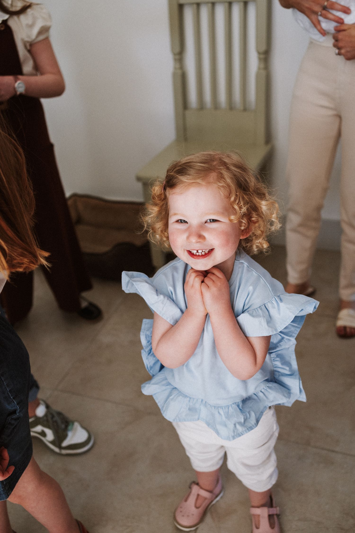 A small child in a light blue ruffled dress and white shorts smiles joyfully while playing indoors.