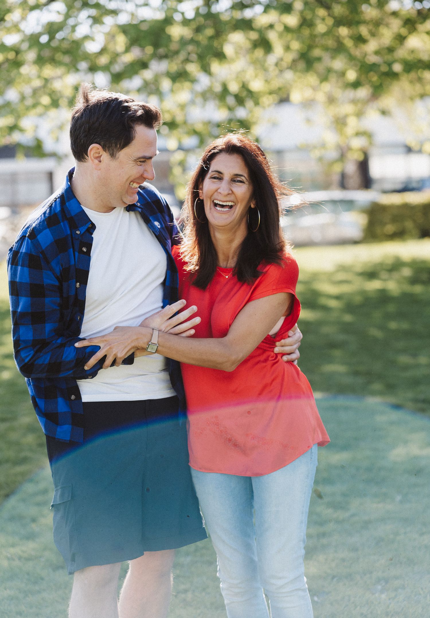 A couple in a blue plaid shirt and red top share playful candid moments outdoors in a grassy park setting.