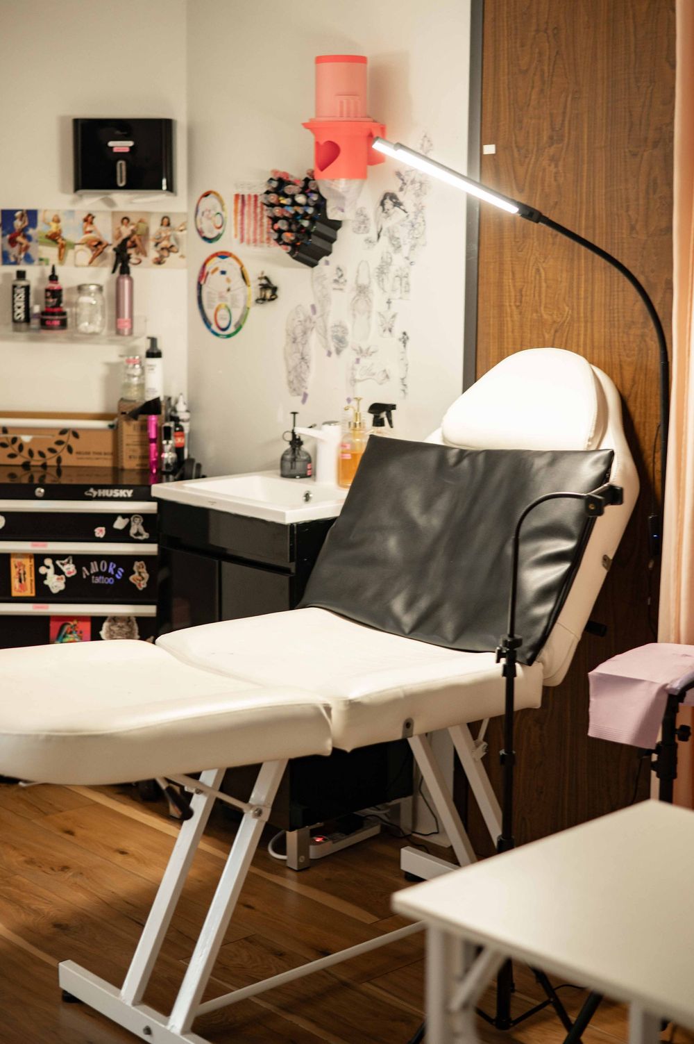 Professional tattoo studio setup with white reclining chair, adjustable lamp, and decorated wall in a cozy wooden interior.