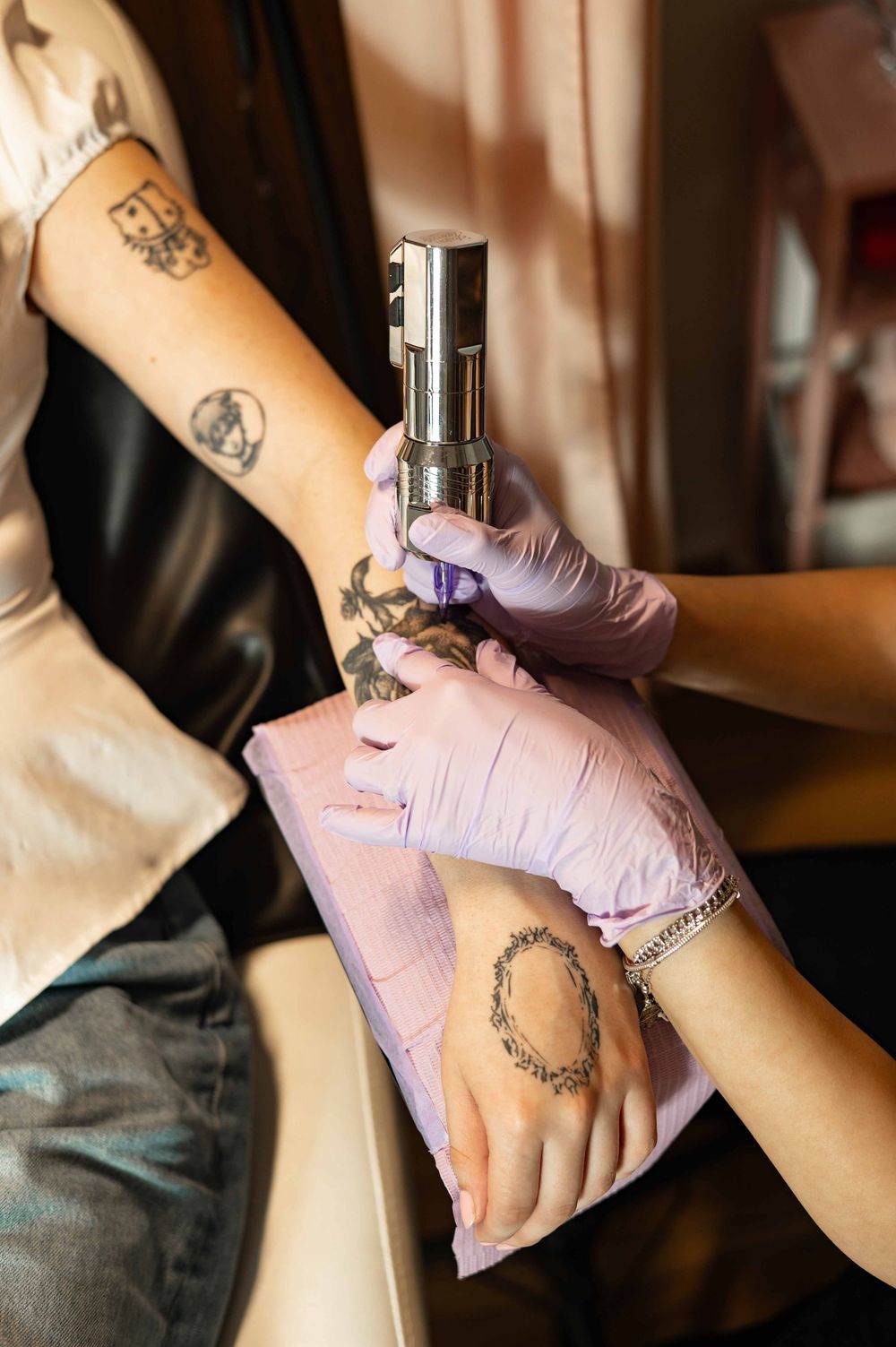 Close-up of a tattoo artist wearing purple gloves working on a client's arm with visible existing tattoos.