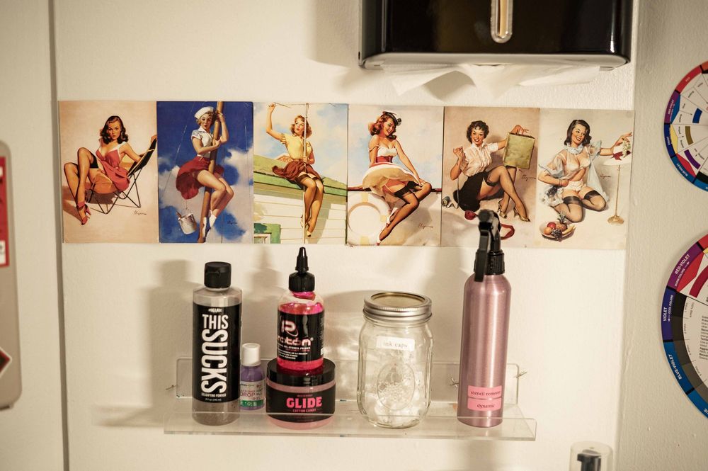 Vintage pin-up posters decorate a shelf with tattoo supplies and ink bottles in a studio setting.