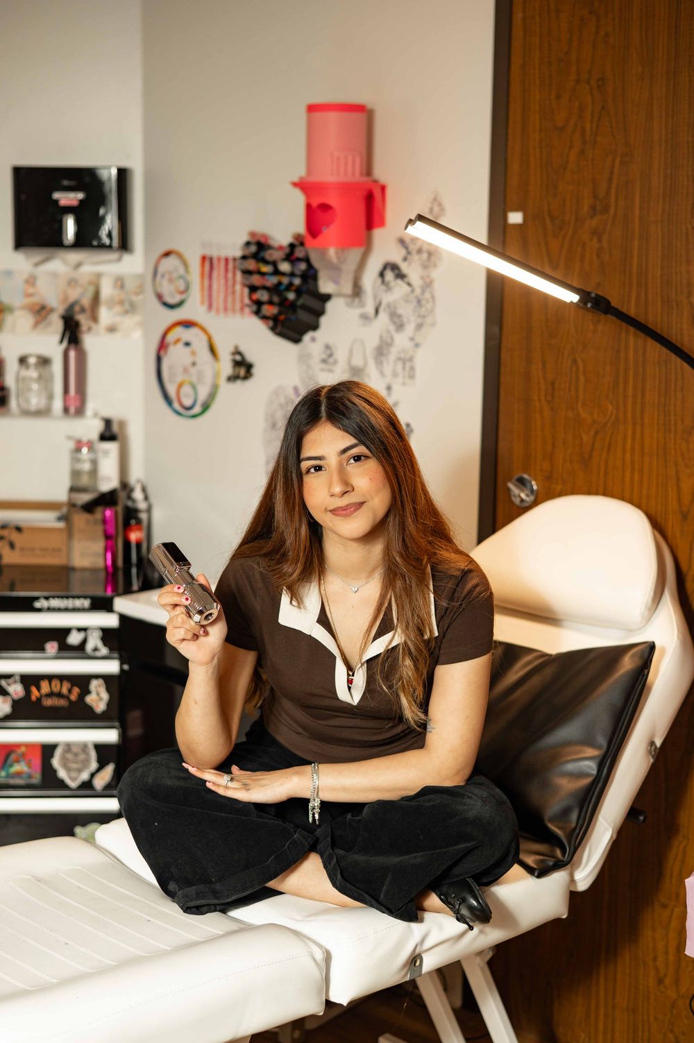 A person relaxes on a white bed while holding a tattoo gun in a casual indoor setting.