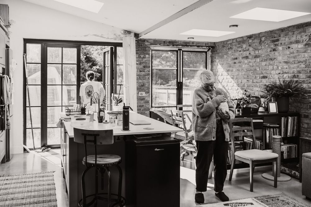 A black and white photo showing a mother with her newborn baby in a living space with exposed brick walls.
