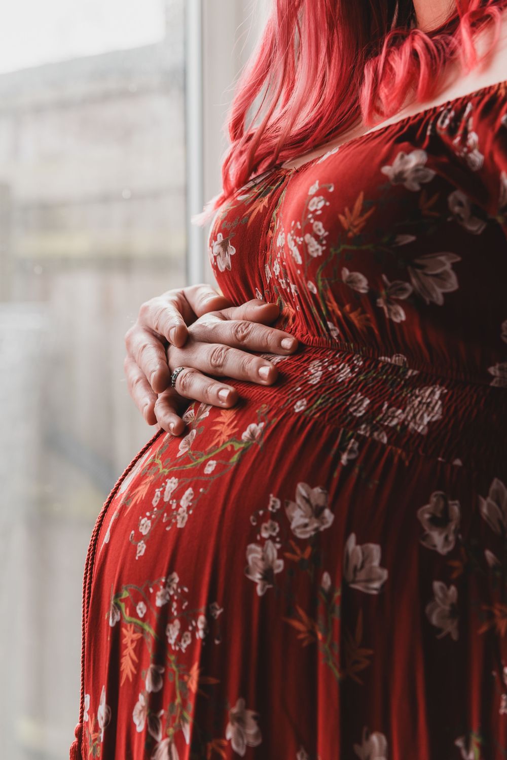 A mother-to-be with vibrant red hair wearing a dark floral dress stands near a window in soft natural lighting.