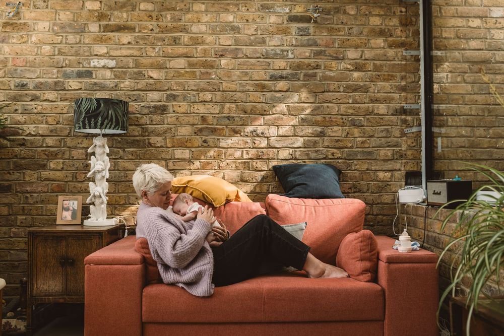A mother and newborn baby relax together on a coral-colored couch against exposed brick wall with table lamp and houseplant.