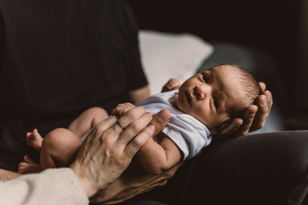 A tiny newborn baby wearing a white onesie rests peacefully on parent's hands in moody natural lighting.