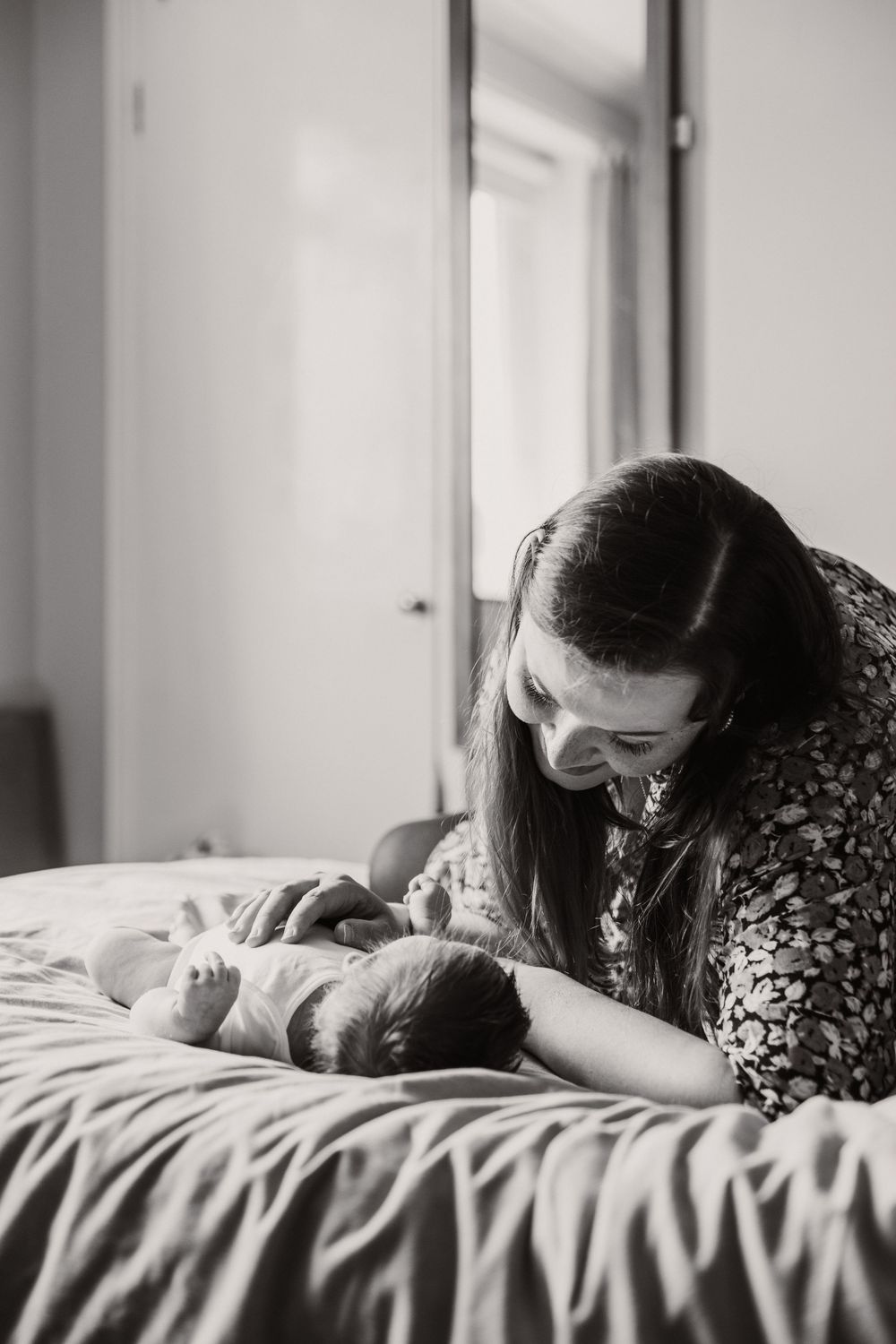A black and white photo showing a tender moment between a mother and newborn baby lying together on a bed.