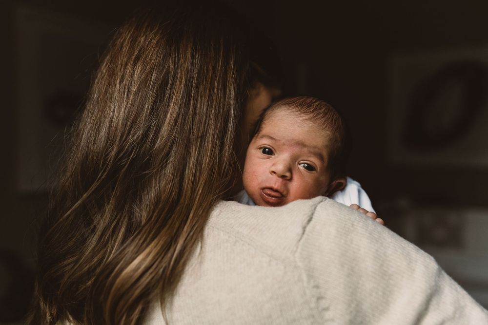 A newborn baby looks straight into the camera over her mother's shoulder in soft window light.