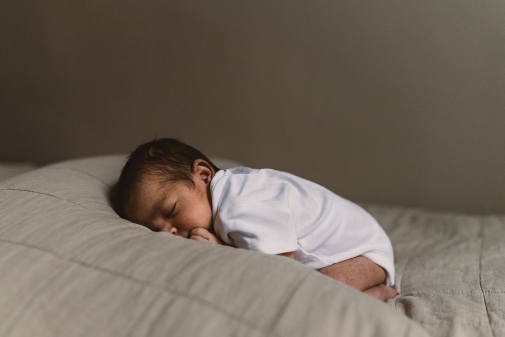 A newborn baby sleeps peacefully on a neutral coloured bed cover with natural light through the window.