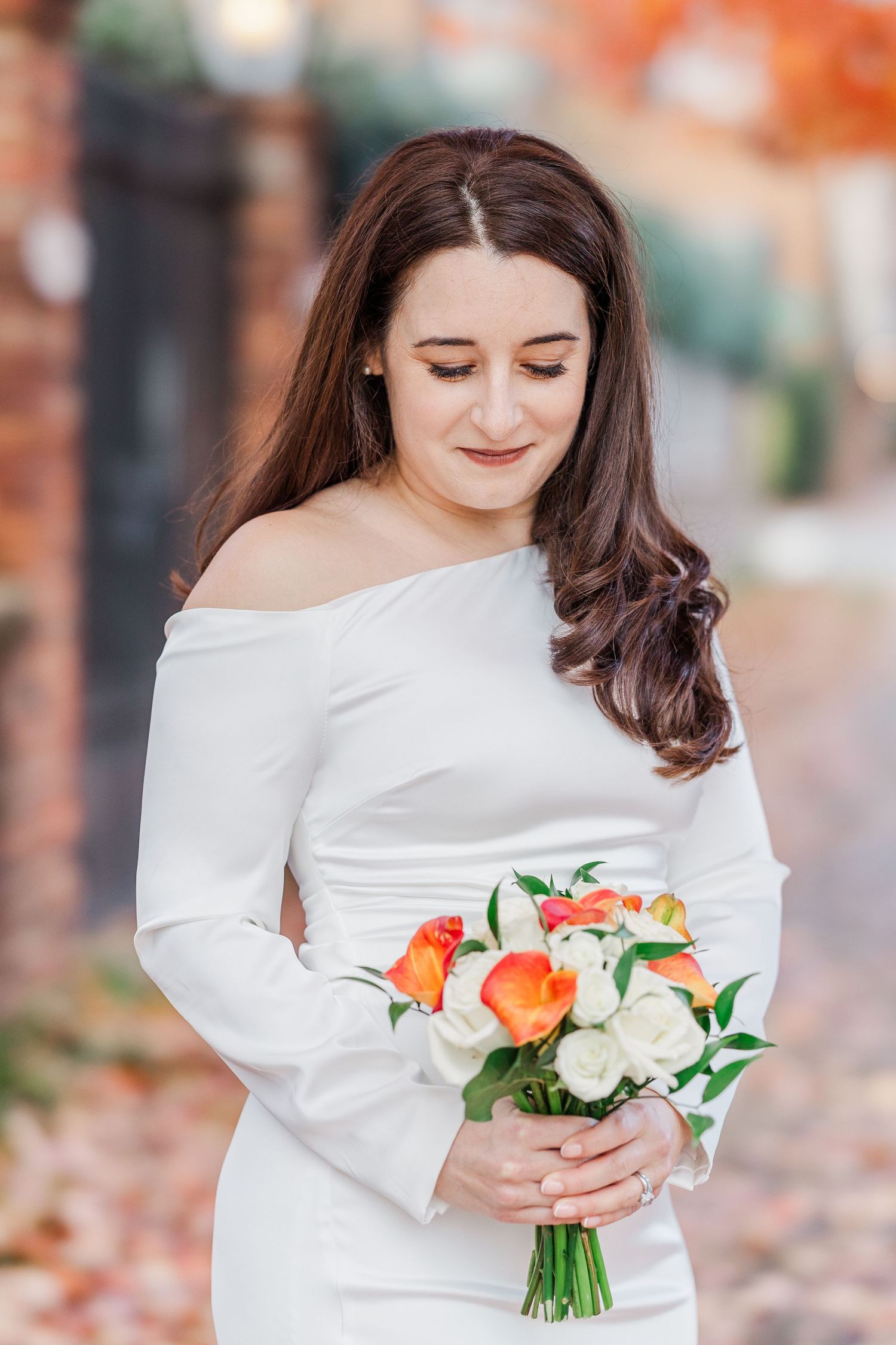 Person in white dress holding orange and white floral bouquet on autumn day with fallen leaves in background.