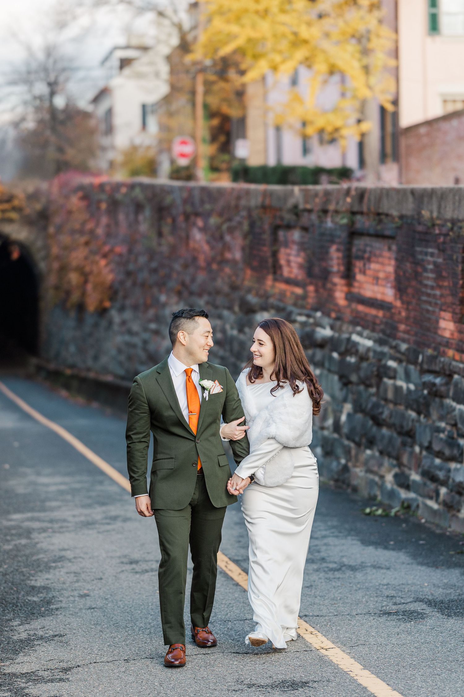 A couple walks along a stone wall path during autumn with fall foliage visible in the background.