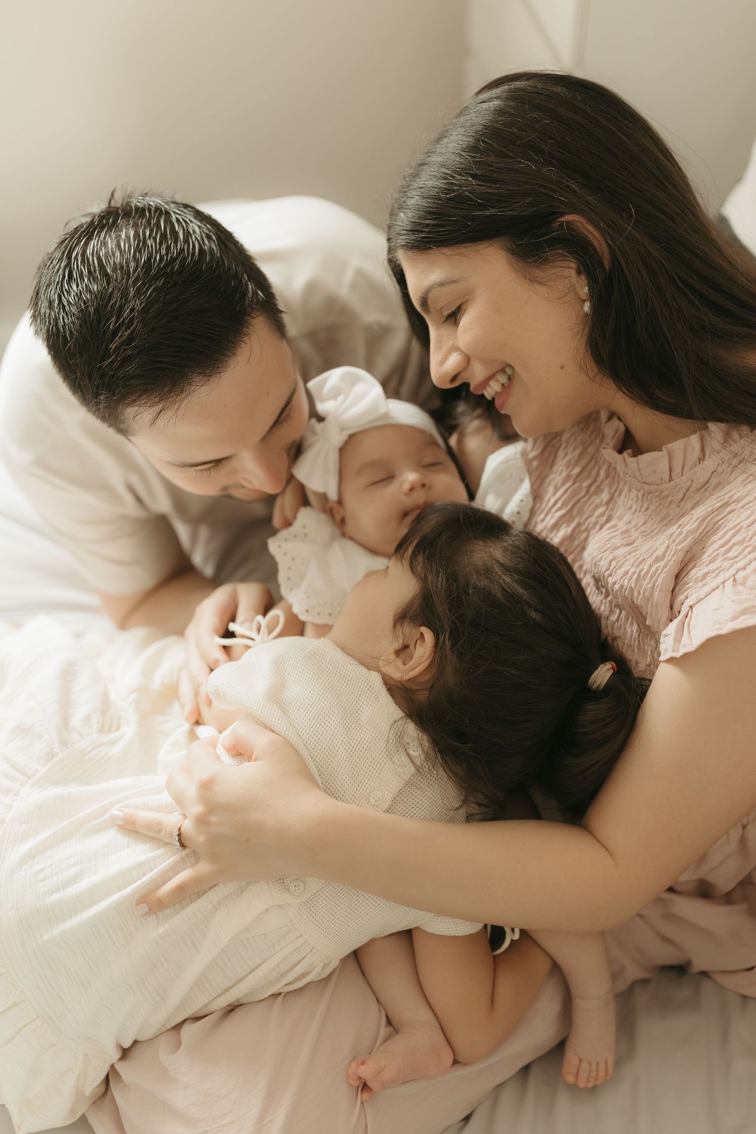 Tender family moment captured in soft light as parents cuddle with their newborn baby on white bedding.