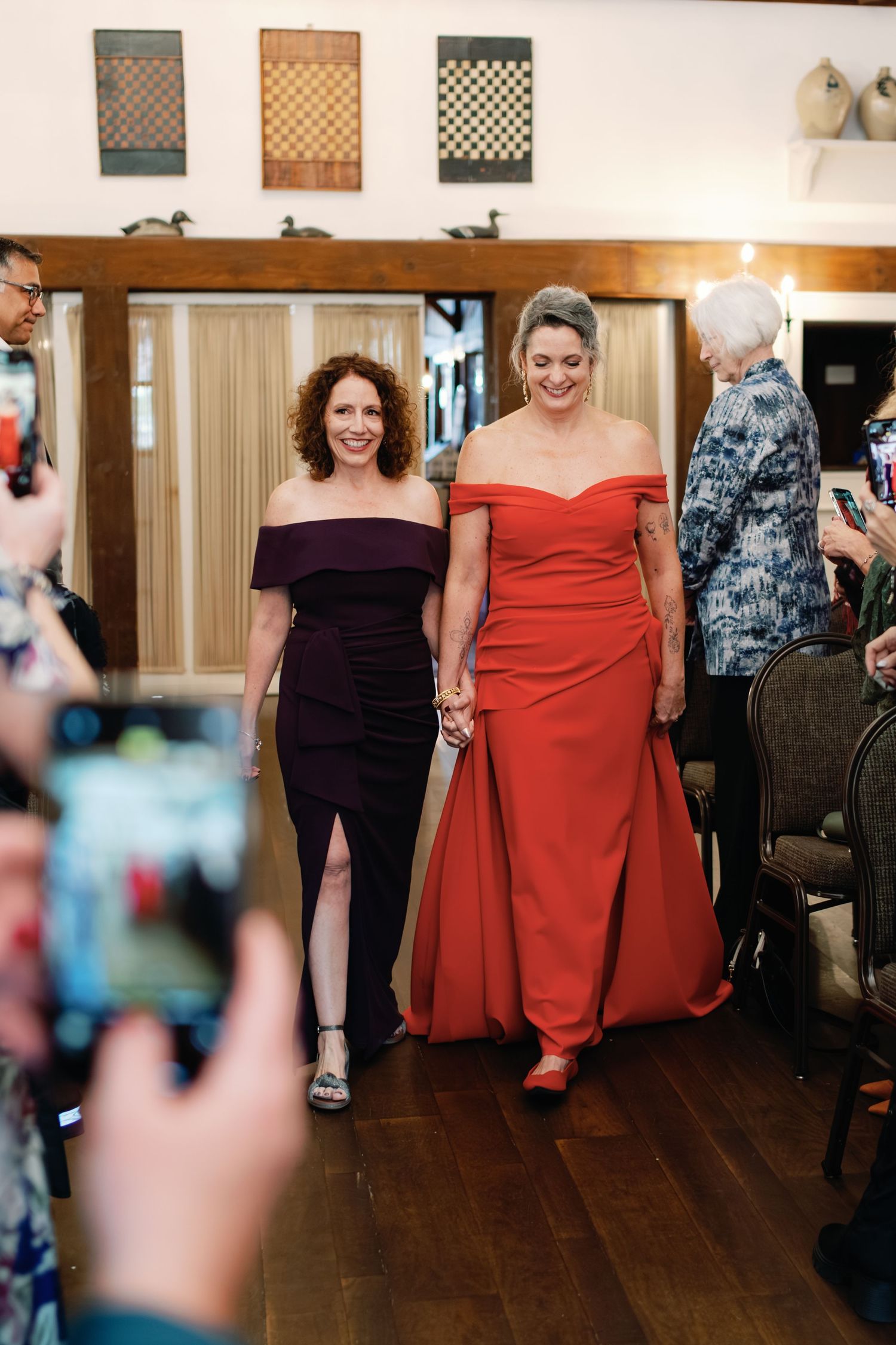 Women in elegant formal dresses pose together at an indoor event with wooden floors and wall decor.