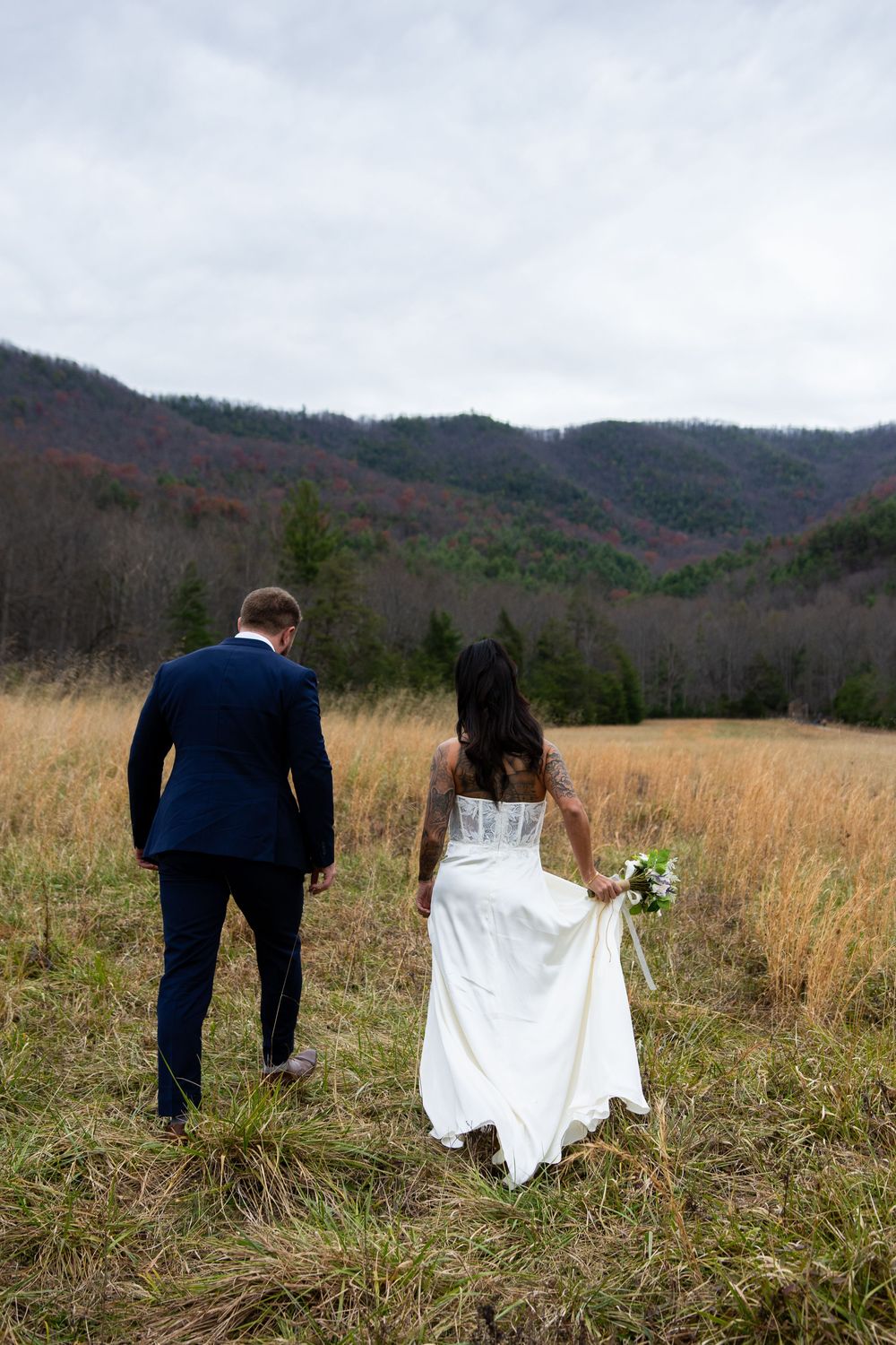 A couple in wedding attire walks through a golden field with mountains in the background during autumn.