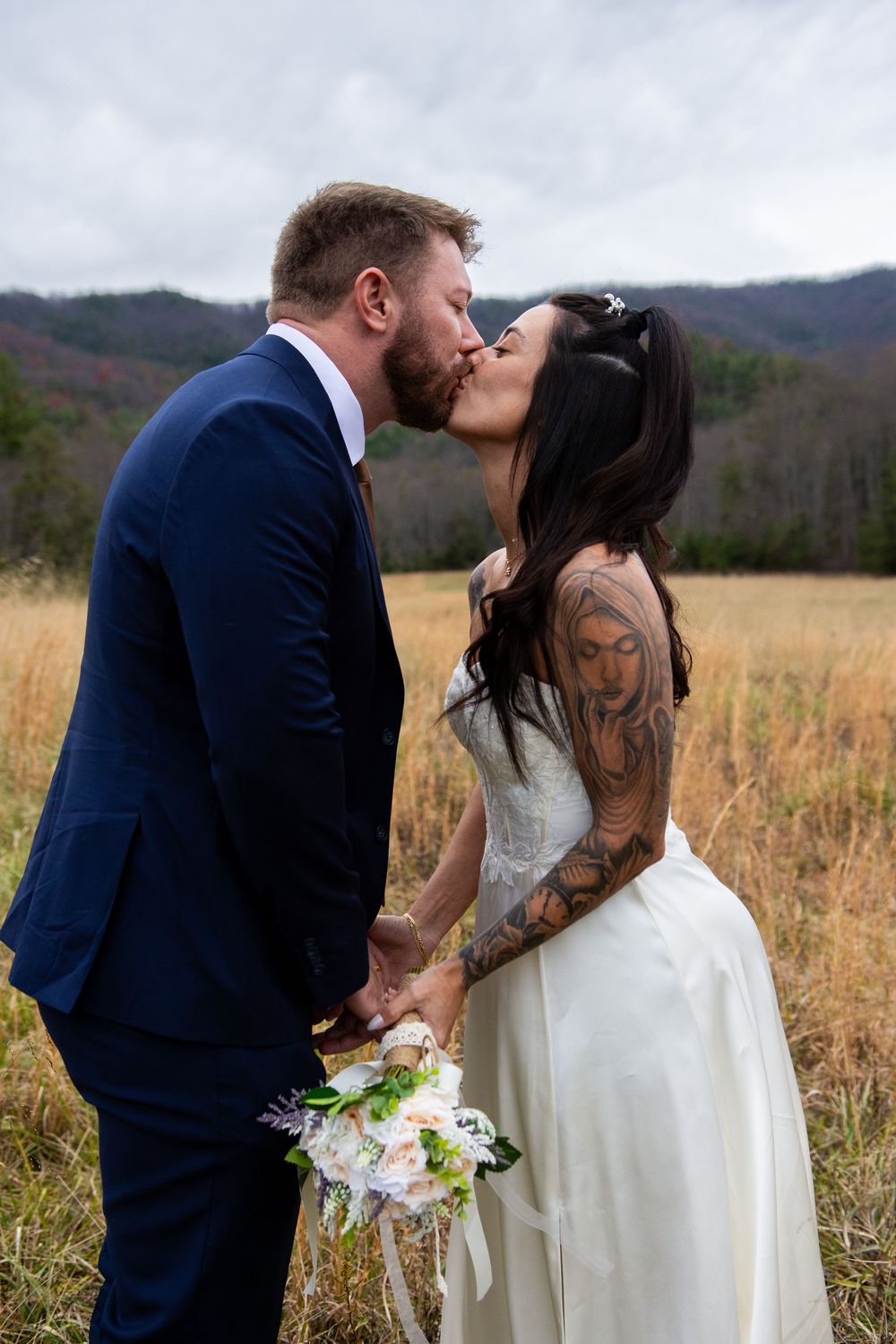 A couple shares a romantic kiss in a golden field with mountains in the background, bride holding a white bouquet.