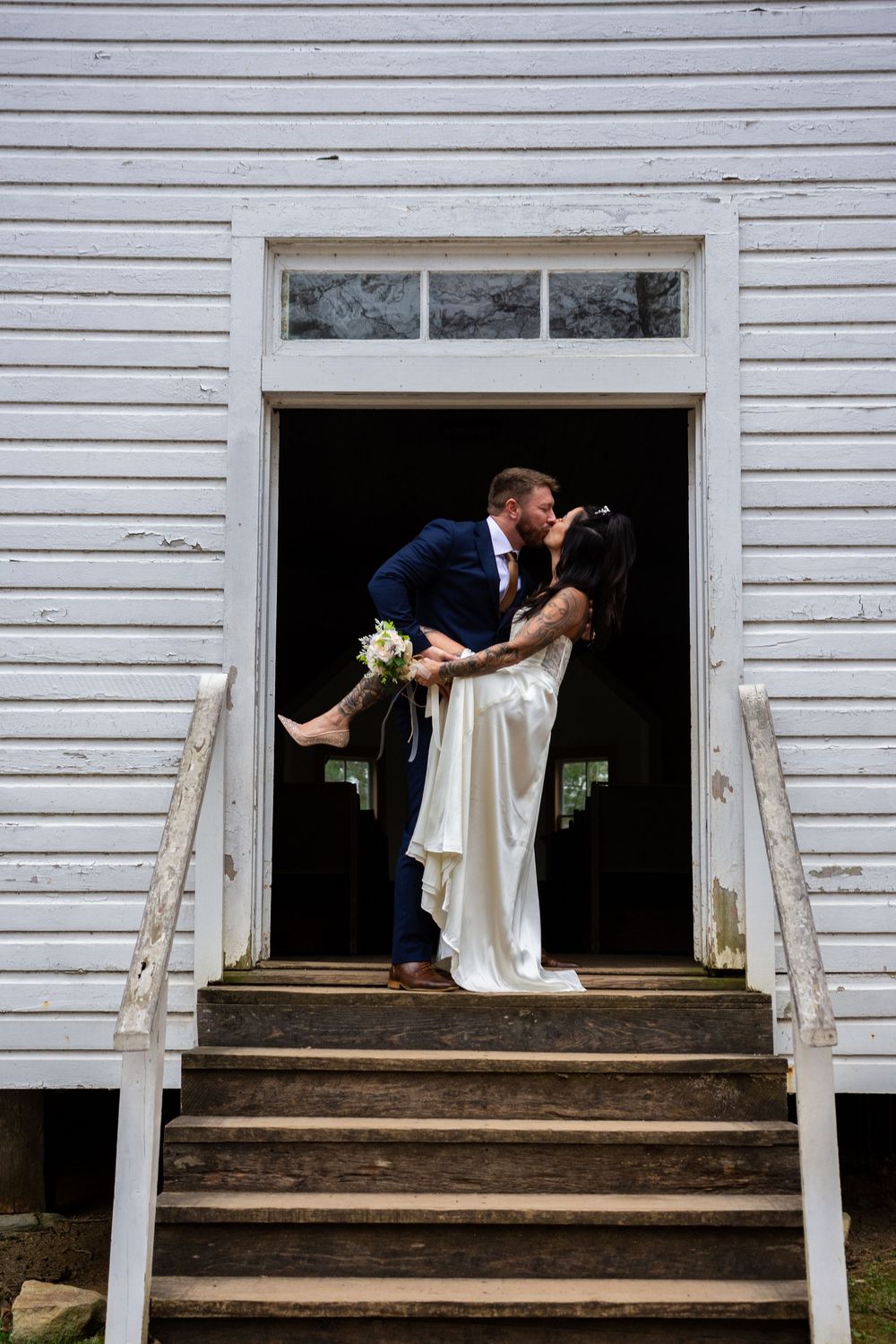 A romantic wedding couple shares a kiss on the steps of a white clapboard church with rustic wooden stairs.