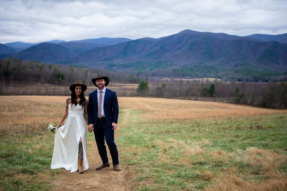 A couple walks through a scenic mountain landscape with rolling hills and autumn foliage in the background.