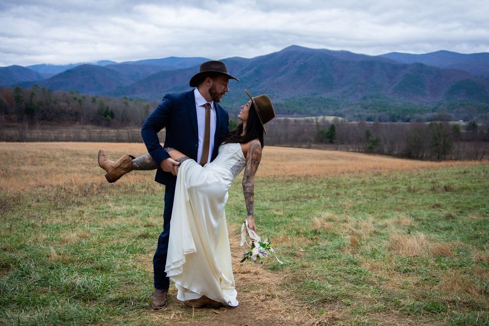 A couple shares a romantic moment in a white dress and suit against a backdrop of rolling mountains.