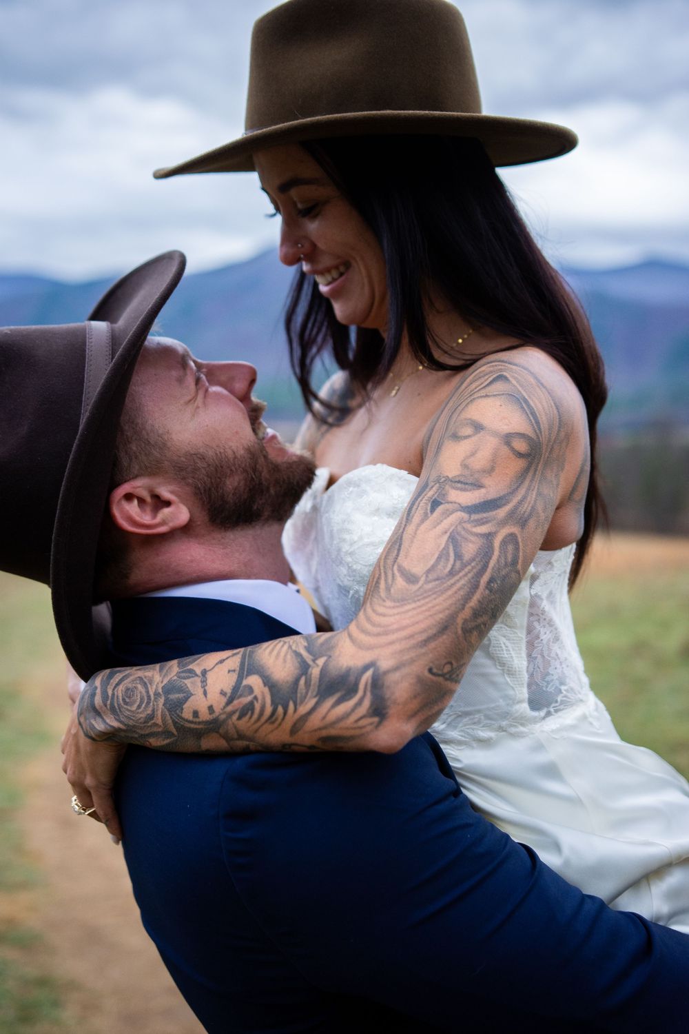 A romantic wedding couple embraces against a mountain backdrop, with the bride in white and groom in navy blue.