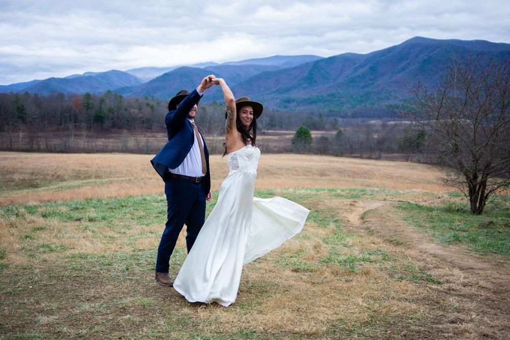 A couple dances joyfully in a field with mountains and rolling hills as their scenic backdrop.