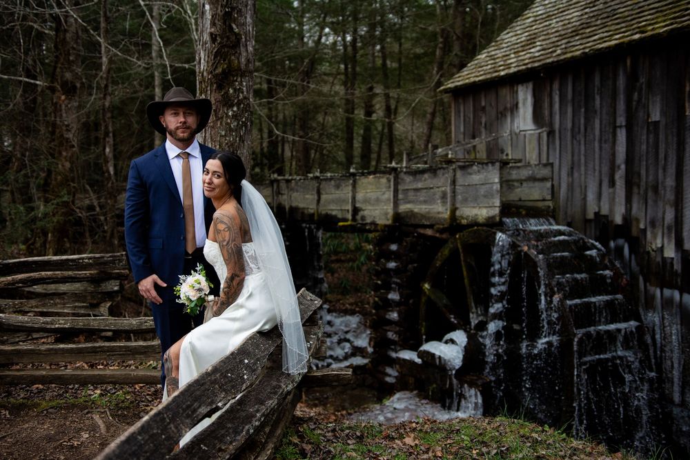 A romantic wedding couple poses together in front of rustic wooden barns and trees in a natural outdoor setting.
