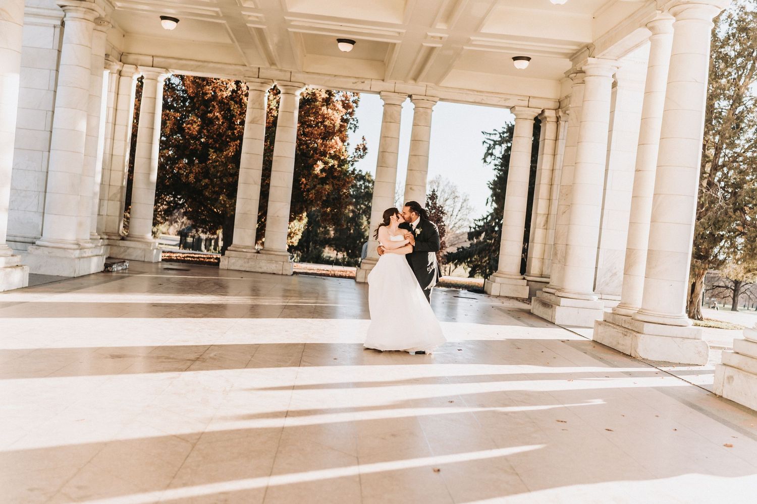 A romantic wedding couple shares a kiss between elegant white columns in a bright outdoor setting.