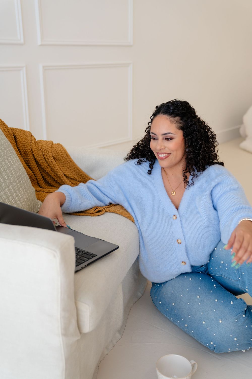 Person in light blue cardigan and jeans relaxes on white couch with laptop and decorative mustard pillow.