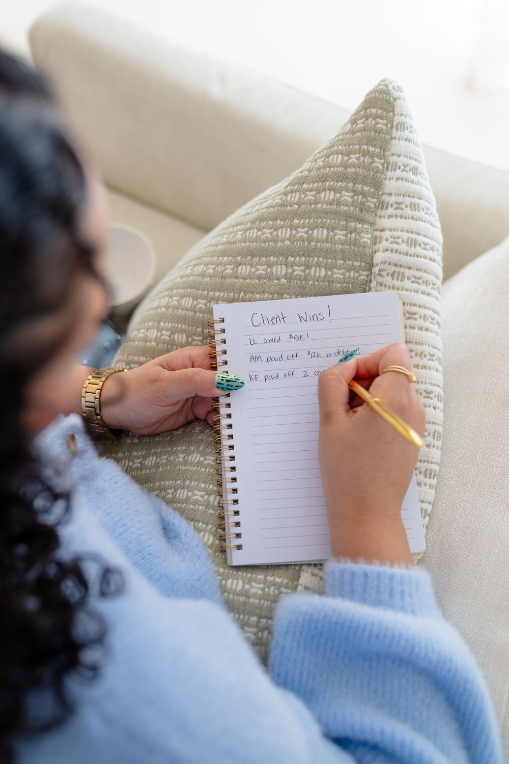 Someone wearing a blue sweater writes in a spiral notebook while resting against a textured pillow.