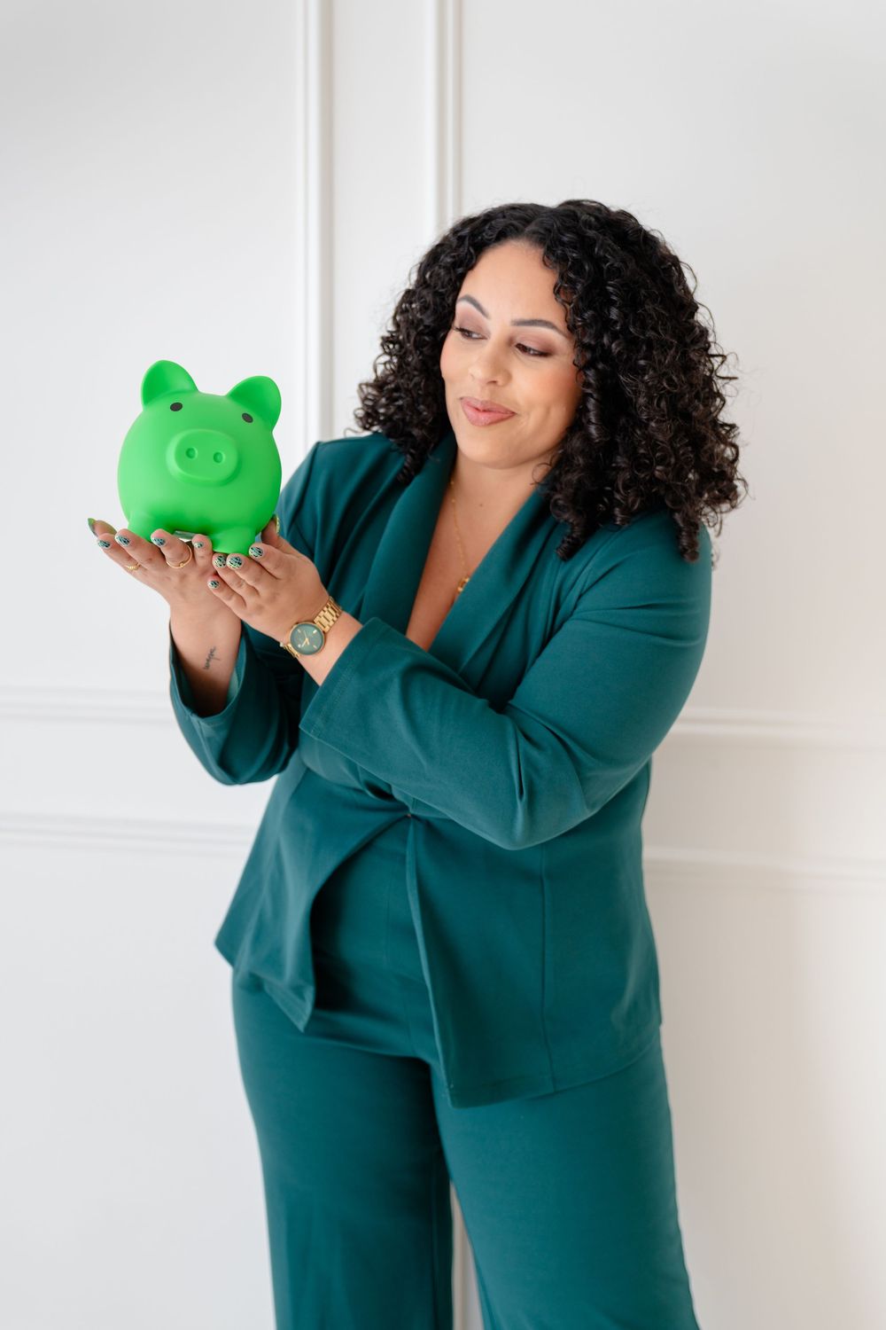 Woman in teal dress poses playfully with green piggy bank against white wall in photo series.
