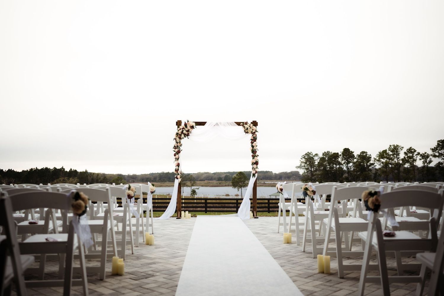 White chairs arranged in rows for an outdoor wedding ceremony with a white aisle runner leading to a floral arch.