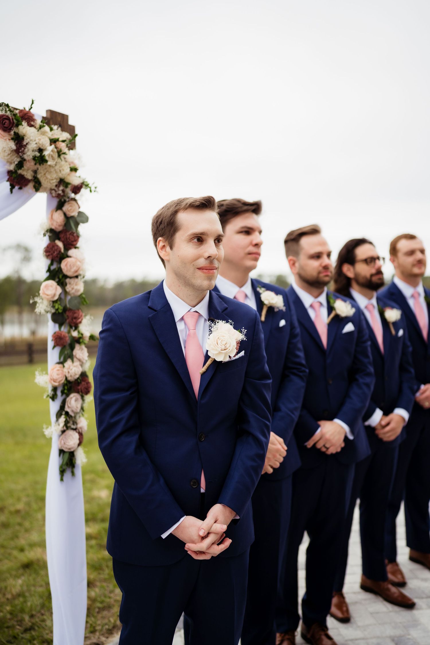 Groomsmen in navy suits with pink ties stand in a line at an outdoor wedding ceremony.