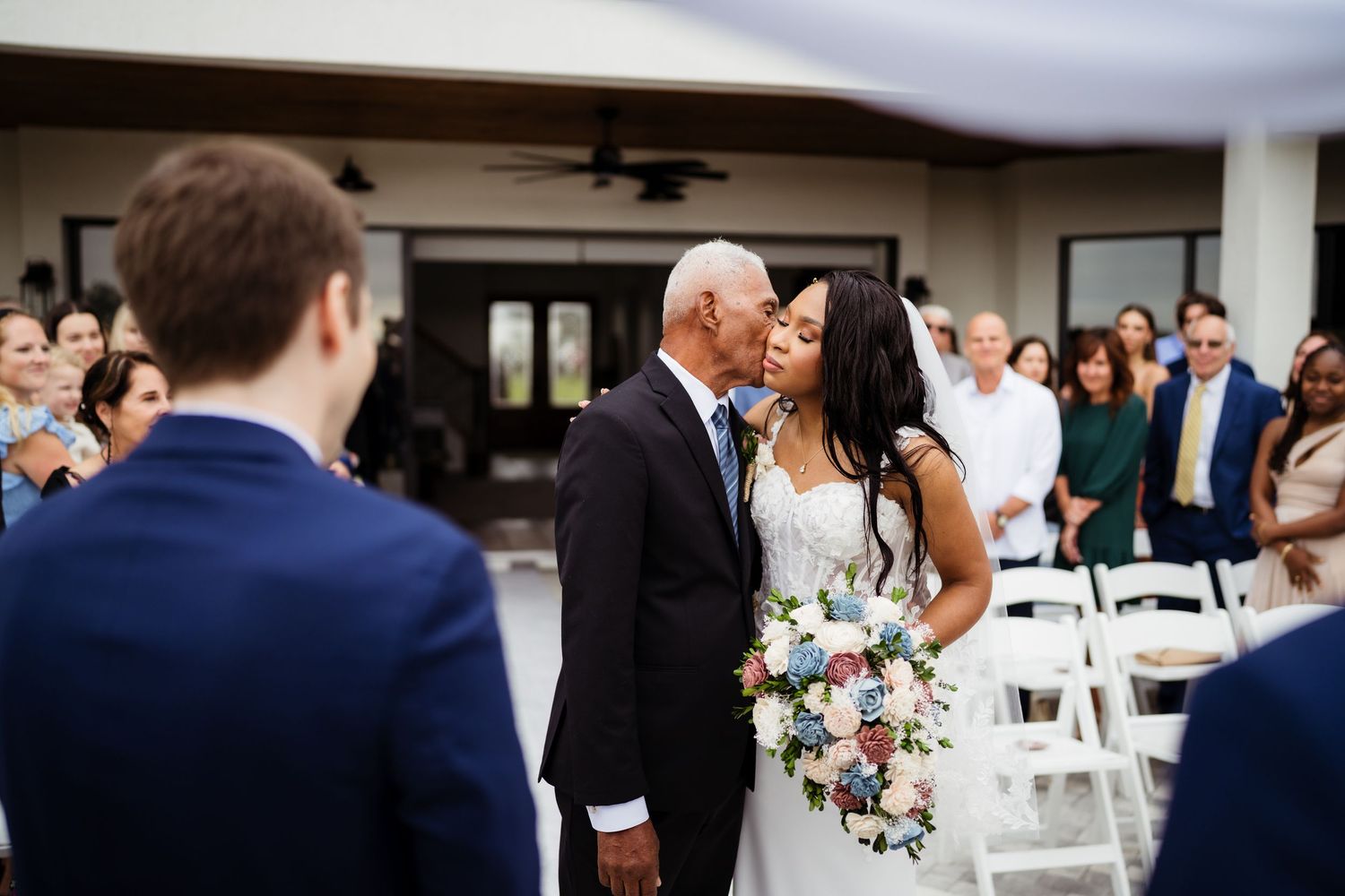 A wedding party gathers around tables outdoors under a covered patio during a reception celebration.